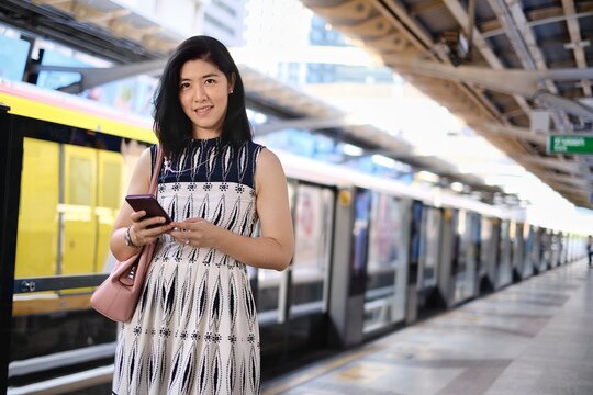 An Attractive Young Confident Asian Business Woman Is Using Her Cellphone While Waiting For Her Train At A Station To Go To Work In The Morning.