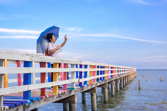 Perspective side view of Asian female tourist with umbrella using smartphone to taking picture on colorful wooden bridge at sea viewpoint against cloud on blue sky background 