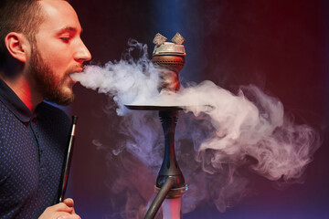 a young man smokes a hookah in an atmospheric hookah room illuminated by color filters and exhales...