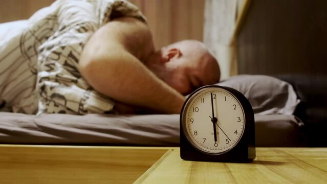 Get up early in the morning on the clock. Sleeping man reluctantly wakes up to an alarm clock early in the morning. The man's hand stops the alarm clock to sleep longer.
