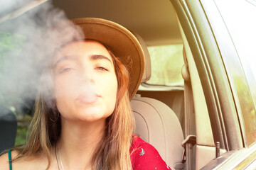 Young woman sitting in a car looking at the camera while blowing smoke from an electronic...
