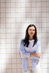brunette woman in blue shirt standing with crossed arms near white tiled wall.