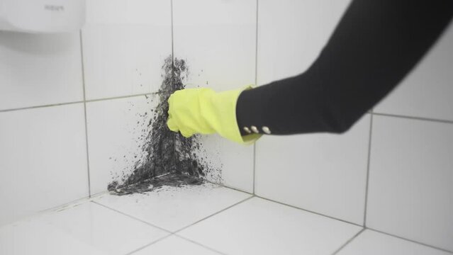 A woman's hand in a yellow rubber glove cleans dirt with a sponge on a white tile