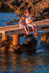 Summer holiday and travel concept. Portrait of young mother and cute daughter sitting on the bridge and meeting the morning sunrise near the sea.