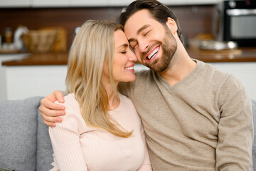 Gentle and lovely couple in love sitting in embrace on the comfortable sofa at home, bonding to each other tenderly. Concept of support and caring in marriage
