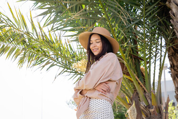 smiling brunette woman in straw hat standing with crossed arms under green palm tree.