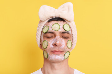 Calm young adult man wearing white t shirt and hair band with bow, posing with face mask and cucumber, standing with closed eyes, relaxing on spa procedures, isolated over yellow background.