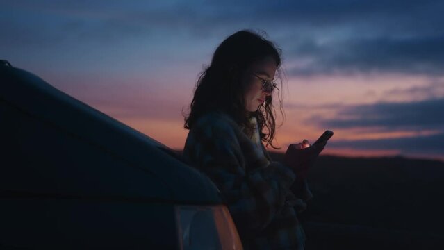 Cinematic lens view soft vintage focus on young pretty woman wearing eyeglasses, watching at phone, chatting with friends, texting, scrolling social media news with colorful stunning sunset background