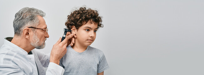 Positive curly male child while ear examining with audiologist. ENT doctor checking boy ear using otoscope - obrazy, fototapety, plakaty
