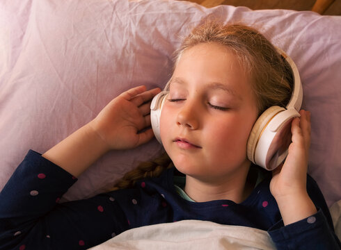 Blonde Child Girl In Headphones Listening To Music For Better Sleep Relaxing Calming Podcast For Stress Relief With Smartphone Online In Bed At Home. Audio Book Tales Against Insomnia For Tired Kids.