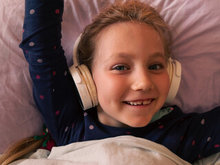 Blonde child girl in headphones learning language listening to music podcast with smartphone online...