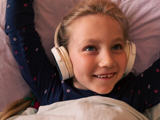 Blonde child girl in headphones learning language listening to music podcast with smartphone online...