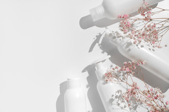 White clean cosmetic bottles, pink dry twigs of flowers on white background flat lay top view. Plastic packaging cosmetic products for branding and label, mock up. Spa, Shampoo, lotion, moisturizer