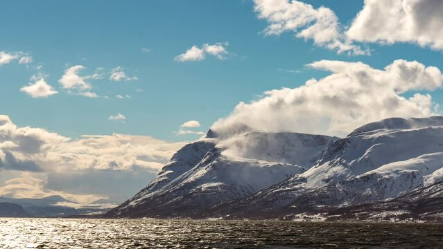 Strong wind blows snow off mountain peak, clouds fly over fjord; time lapse