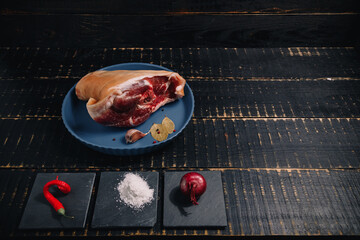 Raw pork knuckle on blue plate, seasonings, garlic, red onions, pepper set, bay leaf, salt on black wooden background. Cooking of meat dish, meal and flavourings, cuisine.
