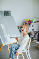 Fototapeta na wymiar Young girl in white shirt and jeans painting picture stands on the easel at home