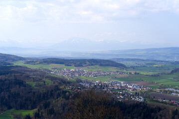 Panoramic view from local mountain Uetliberg with valley, village, agricultural fields and Swiss Alps in the background on a blue cloudy spring day. Photo taken April 14th, 2022, Zurich, Switzerland.