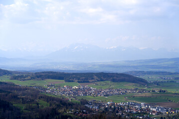 Panoramic view from local mountain Uetliberg with valley, village, agricultural fields and Swiss Alps in the background on a blue cloudy spring day. Photo taken April 14th, 2022, Zurich, Switzerland.
