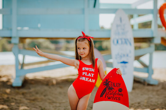 Pretty little girl in red bikini posing with small surfboard like a model on the beach against blue lifeguard tower