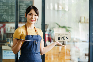 Business owner happy beautiful young asian woman in apron looking at camera, we're open sign on front door smiling welcoming clients to new cafe, People and start-up concept.