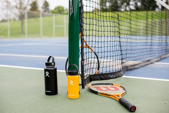 St. Joseph, MO / United States of America - May 24th, 2020 : Pair of Hydro Flask water bottles near tennis court net post, with Wilson racquets lying about.  Tennis court with equipment and hydration.