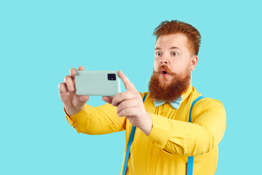 Funny fat man in funky outfit takes selfie on his modern mobile phone. Plus size bearded guy in yellow shirt with bowtie and suspenders recording himself isolated on bright turquoise studio background