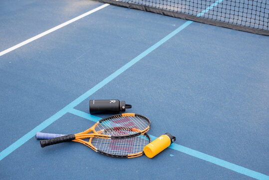 St. Joseph, MO / United States of America - May 24th, 2020 : Pair of tennis rackets and Hydro Flask metal water bottles on tennis court floor.  Stainless steel insulated water bottles and rackets.