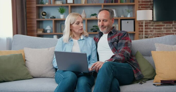 Happy middle aged couple looking at laptop screen sitting on sofa at home. Woman and man in living room searching internet typing online using computer watching video surfing internet.