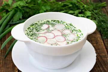 Okroshka in a white deep plate on a wooden table against a background of fresh herbs. Traditional dish of Russian cuisine, summer, cold soup of meat, vegetables and herbs, soft focus.