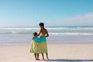 Rear view of african american young mother and daughter with towel standing at beach against sky