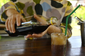 The black soft drink is poured from a plastic bottle into a glass of ice have green straw....