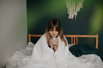 Pretty young woman sits in bed wrapped in a blanket with a cup of coffee