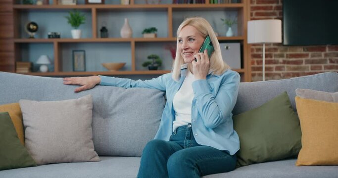 Happy middle aged woman spend leisure alone at home relaxing on sofa, talking on smartphone. Blond famele at home make telephone call talk enjoy conversation.
