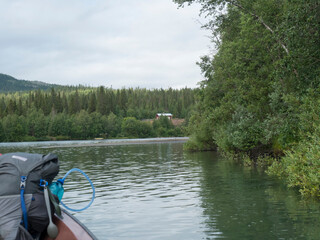 Fototapeta premium View of wide calm Tarra river from boat at Kvikkjokk, with forest and grass shore and green hills. Northern landscape in Swedish Lapland. End of Padjelantaleden hiking trail. Cloudy summer day