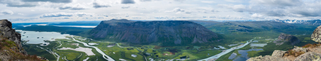 Aerial ultra wide panoramic scenic view from Skierffe rock summit, glacial Rapadalen river delta valley at Sarek national park with meanders, mountains, birch trees. Summer landscape Sweden Lapland - 504392223