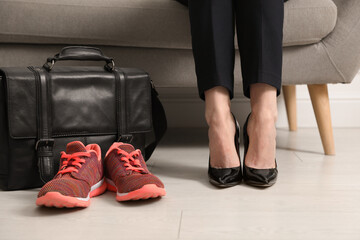 Woman in high heel shoes sitting on sofa near comfortable sneakers in office, closeup