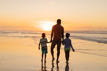 Fototapeta premium Rear view of african american young father holding son and daughter's hands while walking on beach