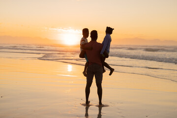 Rear view of african american young man picking up son and daughter on beach against sky at sunset
