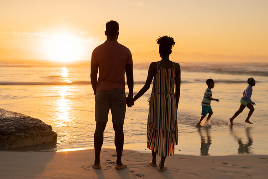 Rear view of african american young couple looking at playful children running on beach against sky - Powered by Adobe