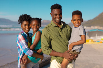 Portrait of african american smiling young parents carrying son and daughter at beach against sky