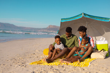 African american young parents with children sitting under umbrella on blanket at beach against sky