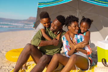 African american boy and girl embracing happy parents sitting on blanket under umbrella at beach