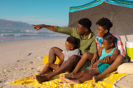 African american young man pointing while sitting with wife and children on blanket against sky