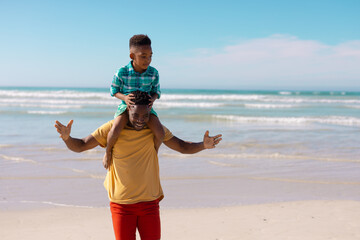 Happy african american young man carrying son on shoulders while standing against sea and sky