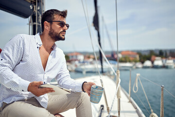 Fototapeta na wymiar A young man with a tablet is sitting and posing for a photo on the yacht while enjoying a coffee and beautiful scenery of the dock on the seaside. Summer, sea, vacation