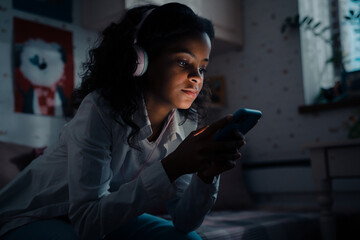 African teen girl with phone listening music and watching video on bed in dark room at home