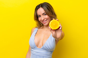 Young caucasian woman isolated on yellow background in swimsuit and holding an orange