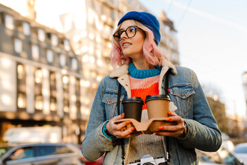 Young woman in eyeglasses holding coffee cups on city street
