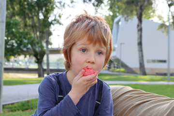 A blond boy is eating a watermelon and the juice is flowing down his arm. A five-year-old boy looks into the distance with interest.