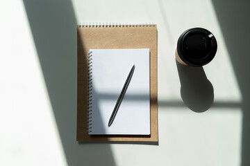 a glass of coffee and a notepad with a pen. craft notepad with pen and cappuccino in sunlight on a white background with beautiful shadows.